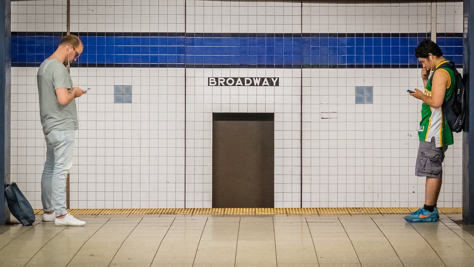 Two commuters on their phones at the Broadway subway platform